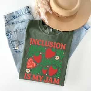 May include: An olive green t-shirt with the text "INCLUSION IS MY JAM" in red. The shirt features a strawberry and leaf design. Light blue jeans, a gold necklace, and a tan hat are also in the image.