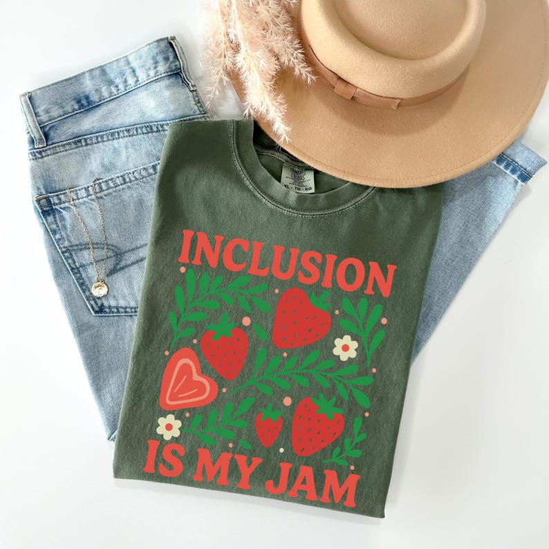 May include: An olive green t-shirt with the text "INCLUSION IS MY JAM" in red. The shirt features a strawberry and leaf design. Light blue jeans, a gold necklace, and a tan hat are also in the image.