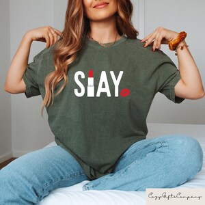 Puede incluir: Camiseta verde oliva con la palabra "SLAY" en letras blancas, un gr&aacute;fico de l&aacute;piz labial rojo y un gr&aacute;fico de labios rojos. La camiseta es de estilo casual.
