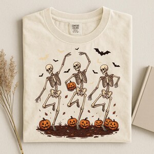 May include: A cream-colored t-shirt featuring a graphic of three skeletons dancing among orange jack-o'-lanterns and black bats. The shirt has a crew neckline and a Comfort Colors brand label printed on the inside of the collar area.