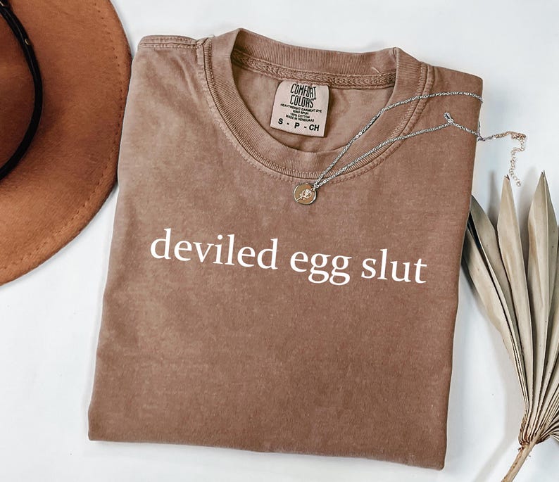 Puede incluir: Una camiseta marr&oacute;n Comfort Colors con la frase "deviled egg slut" impresa en blanco. Un collar plateado con un peque&ntilde;o colgante descansa sobre la camiseta. Un sombrero marr&oacute;n y una planta seca est&aacute;n en el fondo.