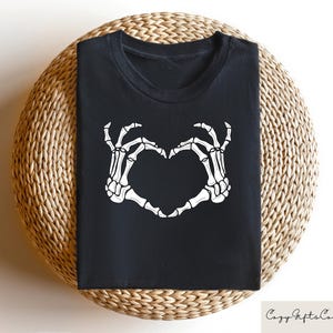 Może przedstawiać: Czarny t-shirt z białym nadrukiem dw&oacute;ch szkieletowych dłoni tworzących kształt serca.