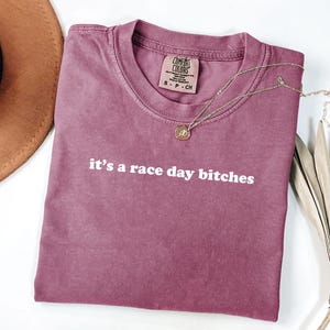 May include: A mauve t-shirt with the white text "it's a race day bitches". A gold necklace with a pendant lies on the shirt. A brown hat and dried plant are in the background. The t-shirt is a small size.