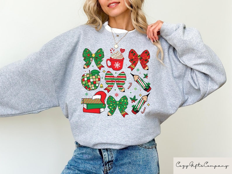 Puede incluir: Sudadera gris con un dise&ntilde;o navide&ntilde;o con lazos rojos y verdes, l&aacute;pices, una taza de chocolate caliente y una manzana.