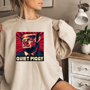 May include: Beige sweatshirt featuring a graphic of a person's face morphed into a pig, set against a red and black radial background. The text "QUIET PIGGY" is displayed below the image in bold, white letters. The sweatshirt has long sleeves.