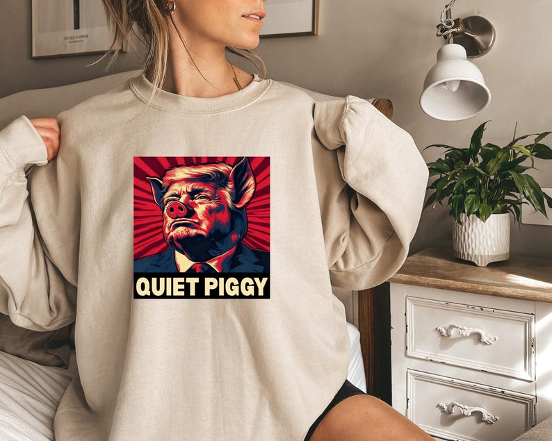 May include: Beige sweatshirt featuring a graphic of a person's face morphed into a pig, set against a red and black radial background. The text "QUIET PIGGY" is displayed below the image in bold, white letters. The sweatshirt has long sleeves.
