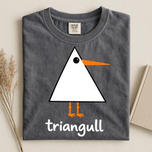 May include: A charcoal gray t-shirt featuring a graphic of a white triangle shaped bird with an orange beak and orange feet. Below the bird, the text reads triangull in white lowercase letters. The shirt has a Comfort Colors brand neck label.
