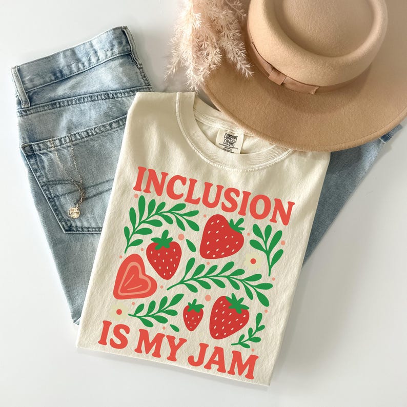 May include: Cream-colored t-shirt with the words "INCLUSION IS MY JAM" in red, along with strawberry and leaf graphics. The shirt is laid on top of blue jeans, a tan hat, and a necklace.