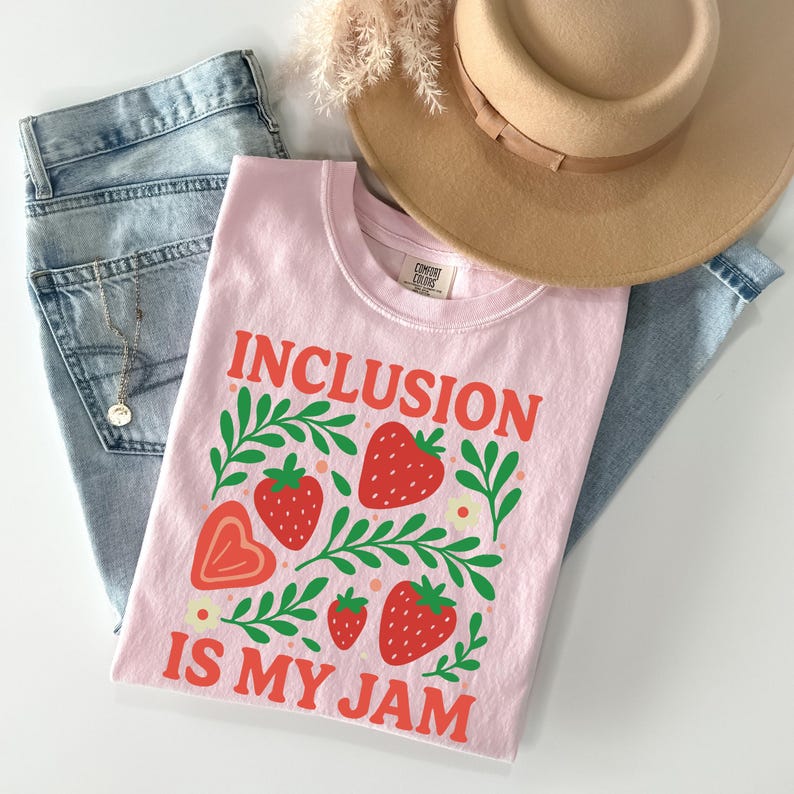 May include: A light pink t-shirt with the words "INCLUSION IS MY JAM" in orange, along with strawberry and leaf graphics. The shirt is paired with light blue jeans and a tan hat. A gold necklace is visible.