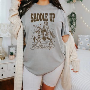 May include: A gray t-shirt with a brown graphic of a woman riding a horse. The graphic says "Saddle Up Buttercup!"