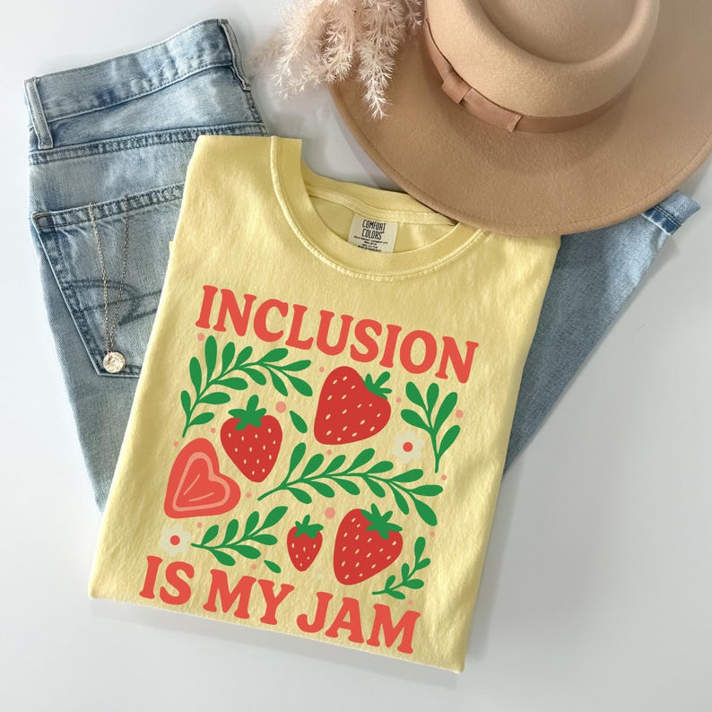 May include: A light yellow t-shirt with the words "INCLUSION IS MY JAM" in red, along with strawberry and leaf graphics. The shirt is folded on top of blue jeans, a necklace, and a tan hat.