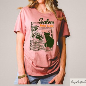 May include: A dusty rose t-shirt featuring a graphic for the "Salem Book Club." The design includes a black cat, a stack of books, a witch, and a haunted house. The text reads "Books, Booze, Black Cats, All Witches Welcome, Every Turning is Saturday."