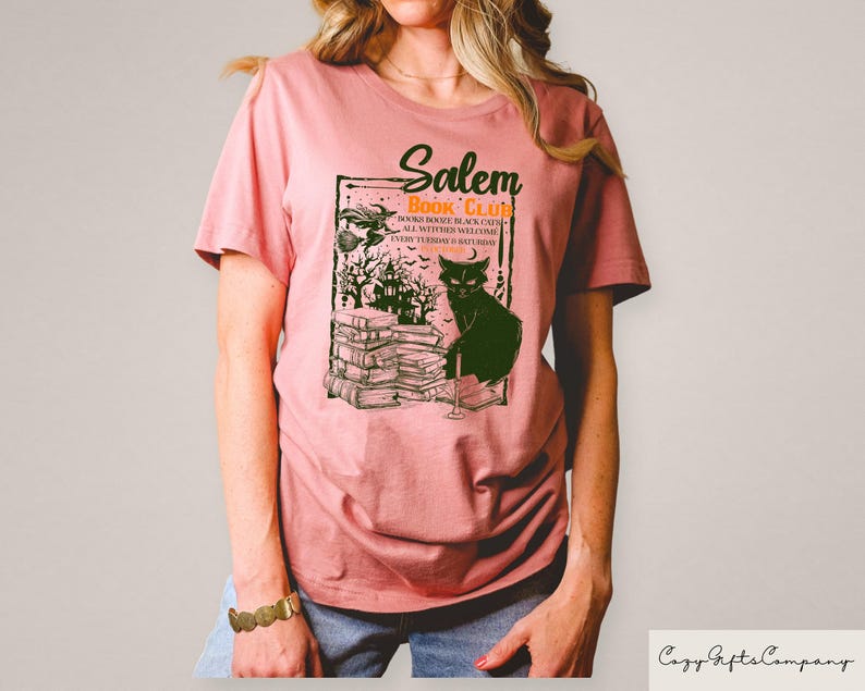 May include: A dusty rose t-shirt featuring a graphic for the "Salem Book Club." The design includes a black cat, a stack of books, a witch, and a haunted house. The text reads "Books, Booze, Black Cats, All Witches Welcome, Every Turning is Saturday."