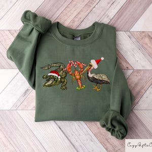 May include: A green sweatshirt with a Christmas-themed design featuring an alligator, a pelican, and a lobster wearing Santa hats. The alligator is holding a candy cane.