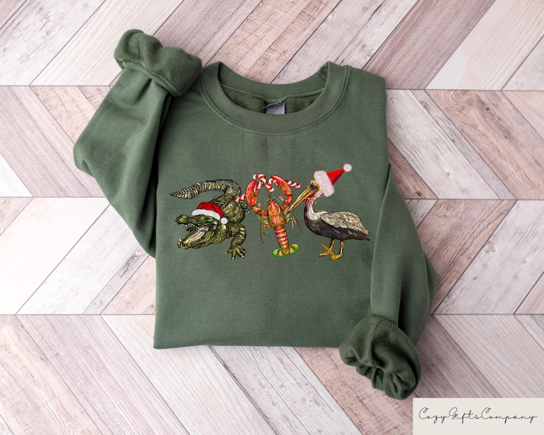 May include: A green sweatshirt with a Christmas-themed design featuring an alligator, a pelican, and a lobster wearing Santa hats. The alligator is holding a candy cane.