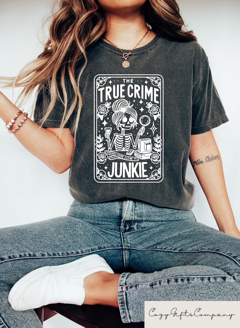May include: A dark grey t-shirt featuring a white graphic of a skeleton holding a glass of wine and a magnifying glass. The graphic is surrounded by roses and stars. The text "THE TRUE CRIME JUNKIE" is printed on the graphic.