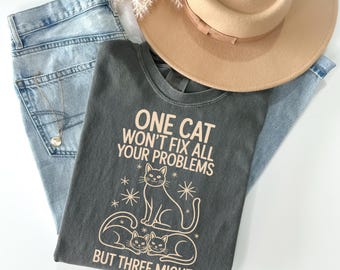 May include: A casual flat lay featuring a gray t-shirt with the text "ONE CAT WON'T FIX ALL YOUR PROBLEMS BUT THREE MIGHT" and cat illustrations. Light blue jeans, a tan hat, and a necklace complete the ensemble.