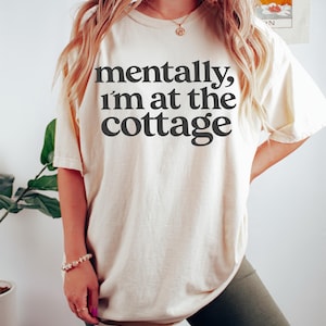 以下が含まれることがあります： 「mentally, I'm at the cottage」の黒い文字がプリントされたクリーム色のTシャツ。ダークグリーンのサイクリングショーツと合わせています。ブレスレットとネックレスが見えます。