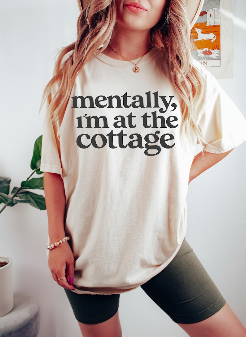 以下が含まれることがあります： 「mentally, I'm at the cottage」の黒い文字がプリントされたクリーム色のTシャツ。ダークグリーンのサイクリングショーツと合わせています。ブレスレットとネックレスが見えます。