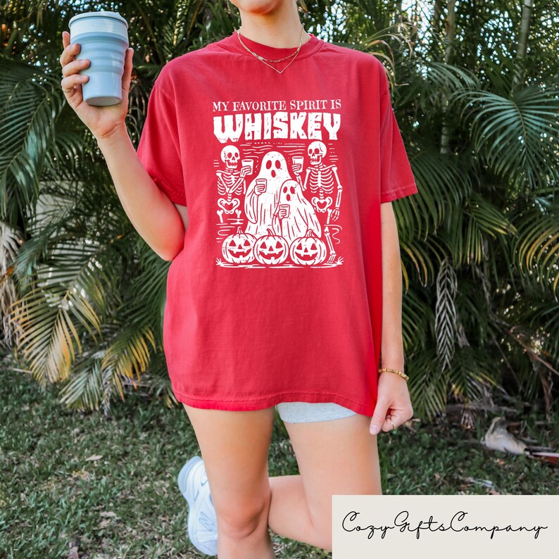 May include: Red t-shirt with the text "MY FAVORITE SPIRIT IS WHISKEY" in white. The graphic includes skeletons, ghosts, and jack-o'-lanterns. The shirt is a Halloween-themed design.