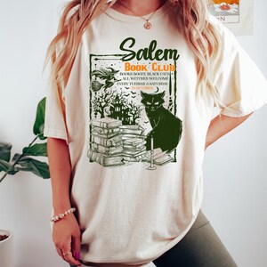 May include: Cream-colored t-shirt with a green graphic featuring the text "Salem Book Club." The design includes a black cat, stacks of books, and a spooky haunted house. The shirt also has the text "Books Booze Black Cats All Witches Welcome Every Tuesday & Saturday in October."