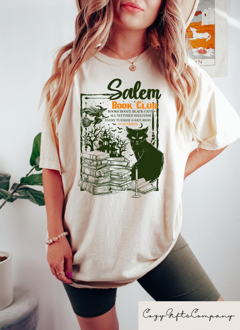 May include: Cream-colored t-shirt with a green graphic featuring the text "Salem Book Club." The design includes a black cat, stacks of books, and a spooky haunted house. The shirt also has the text "Books Booze Black Cats All Witches Welcome Every Tuesday & Saturday in October."