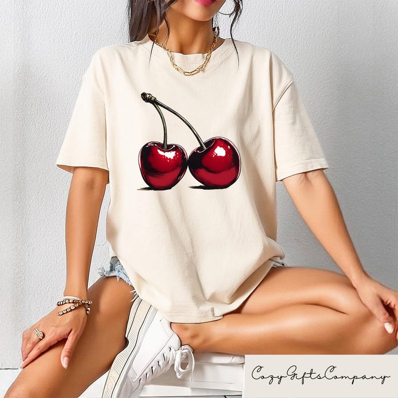 May include: A cream colored t-shirt with a graphic of two red cherries on the front. The cherries have green stems and leaves.