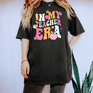May include: Black t-shirt featuring the phrase "IN MY TEACHER ERA" in a vibrant, retro font. The design incorporates a pencil, stars, a smiley face, and a checkered apple. The shirt has a relaxed, oversized fit.