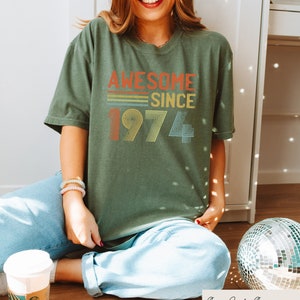 Pu&ograve; includere: Una maglietta verde con il testo "Awesome Since 1974" in un design retr&ograve; arcobaleno.