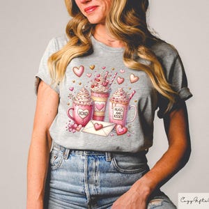 May include: A gray t-shirt featuring a watercolor design of three coffee drinks with whipped cream, hearts, and the phrases "XOXO" and "Hugs and Kisses." A heart-shaped envelope and flowers are also included in the design.