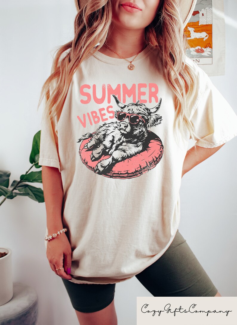 May include: A cream colored t-shirt with a graphic of a cow wearing sunglasses and a pink inflatable ring. The text "SUMMER VIBES" is printed above the cow.