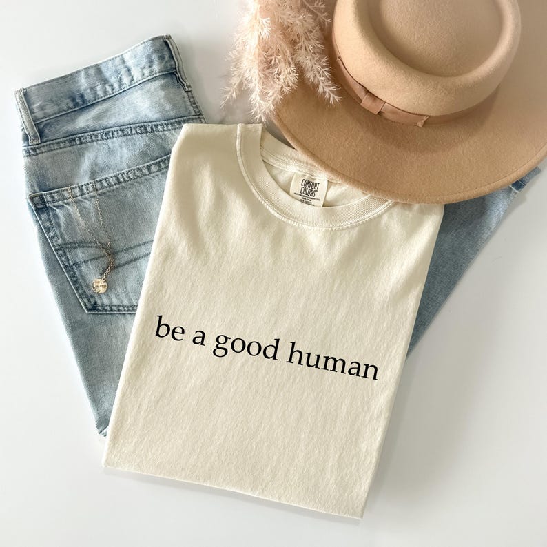 May include: Cream-colored t-shirt with the text "be a good human" in black, paired with light blue denim jeans, a beige hat, and a delicate necklace. The shirt is made of a soft material.