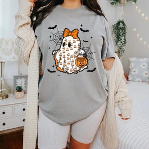 Puede incluir: Una camiseta gris con un dise&ntilde;o de fantasma de Halloween. El fantasma es blanco con detalles en naranja, un lazo y un cubo de caramelos con forma de calabaza. Murci&eacute;lagos negros y telara&ntilde;as rodean al fantasma. El texto "Cozy Gifts Company" est&aacute; en la parte inferior.