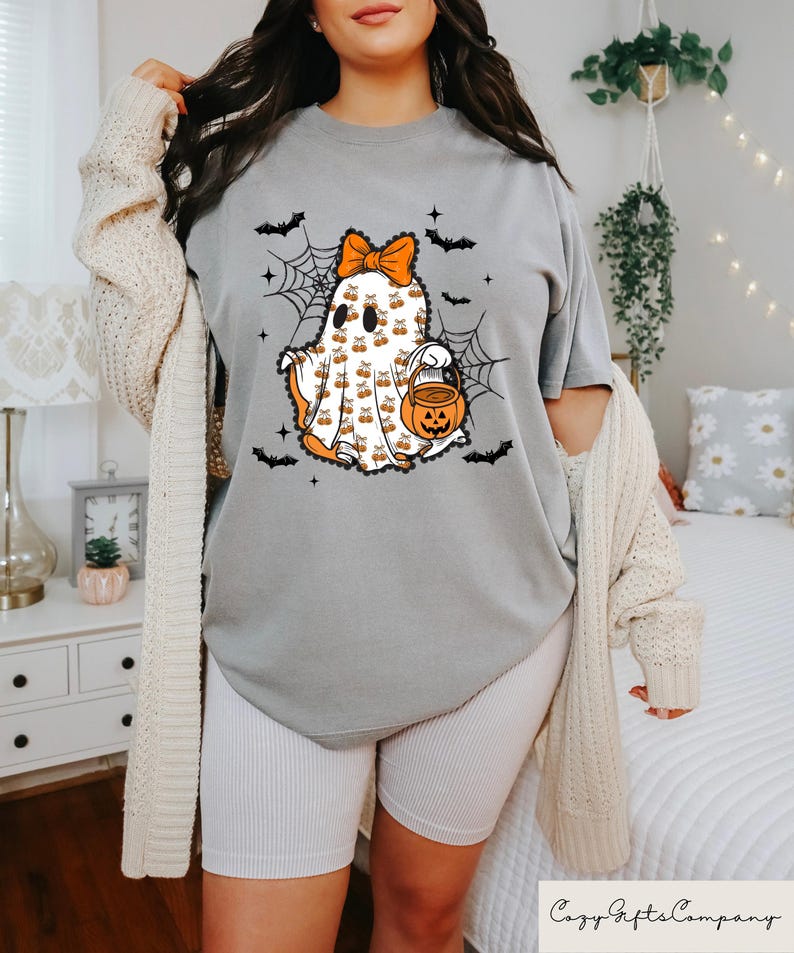 Puede incluir: Una camiseta gris con un dise&ntilde;o de fantasma de Halloween. El fantasma es blanco con detalles en naranja, un lazo y un cubo de caramelos con forma de calabaza. Murci&eacute;lagos negros y telara&ntilde;as rodean al fantasma. El texto "Cozy Gifts Company" est&aacute; en la parte inferior.