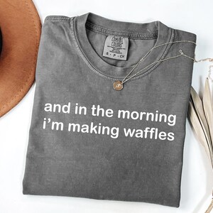 May include: Gray t-shirt with the text "and in the morning I'm making waffles" in white. A gold necklace with a pendant rests on the shirt. A brown hat and dried palm leaves are in the background.