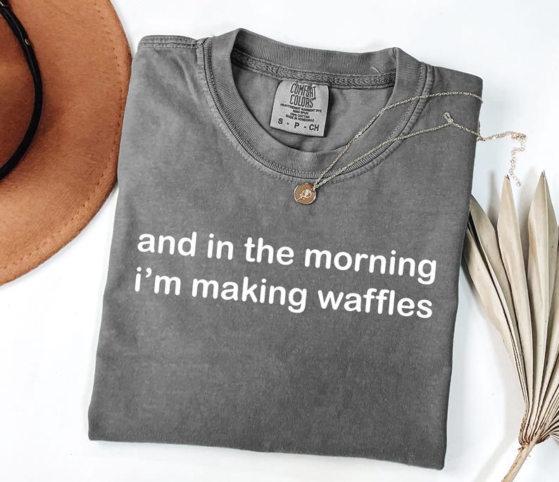 May include: Gray t-shirt with the text "and in the morning I'm making waffles" in white. A gold necklace with a pendant rests on the shirt. A brown hat and dried palm leaves are in the background.