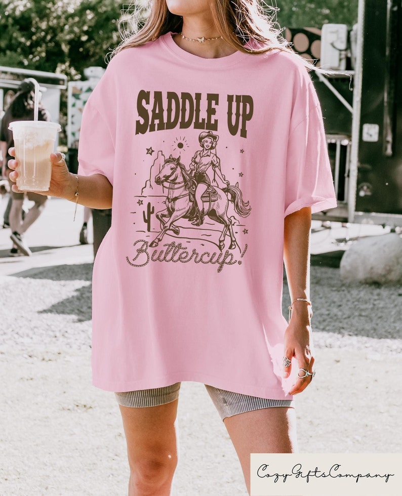 May include: Pink oversized t-shirt with a brown graphic of a woman riding a horse with the text "Saddle Up Buttercup!"