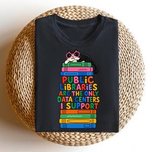 May include: A black t-shirt featuring a graphic of a cat wearing pink glasses resting on a stack of colorful books. The text reads, "PUBLIC LIBRARIES ARE THE ONLY DATA CENTERS I SUPPORT," printed in multi-colored letters between the book stacks.