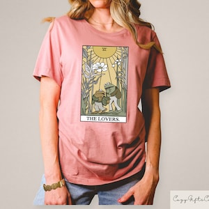 May include: A pink t-shirt featuring a graphic of a tarot card depicting two frogs in front of a flower and sun. The card is labelled "The Lovers."
