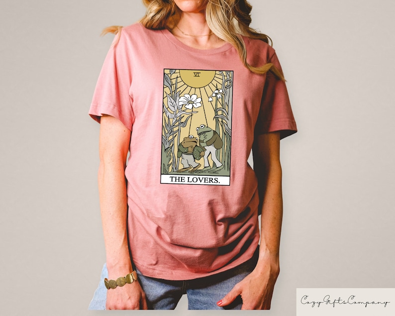 May include: A pink t-shirt featuring a graphic of a tarot card depicting two frogs in front of a flower and sun. The card is labelled "The Lovers."