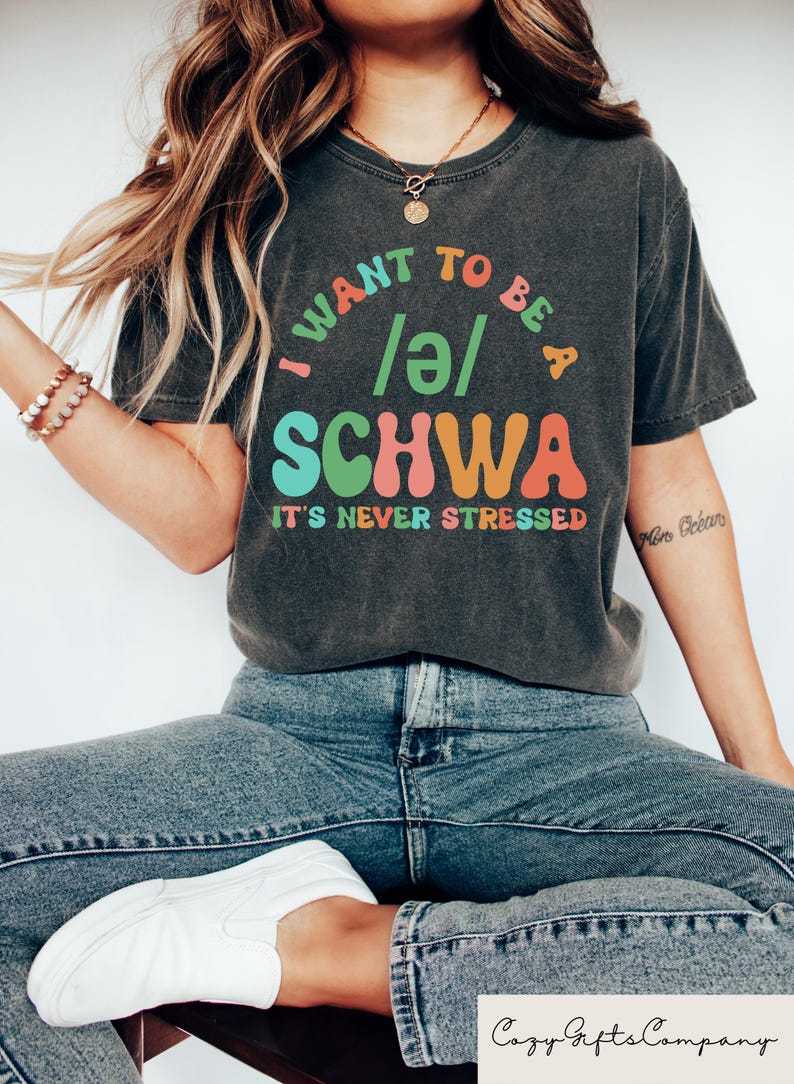 May include: A dark gray t-shirt with the text "I want to be a /ə/ SCHWA it's never stressed" in colorful letters. The shirt is being worn by a person with long blonde hair.