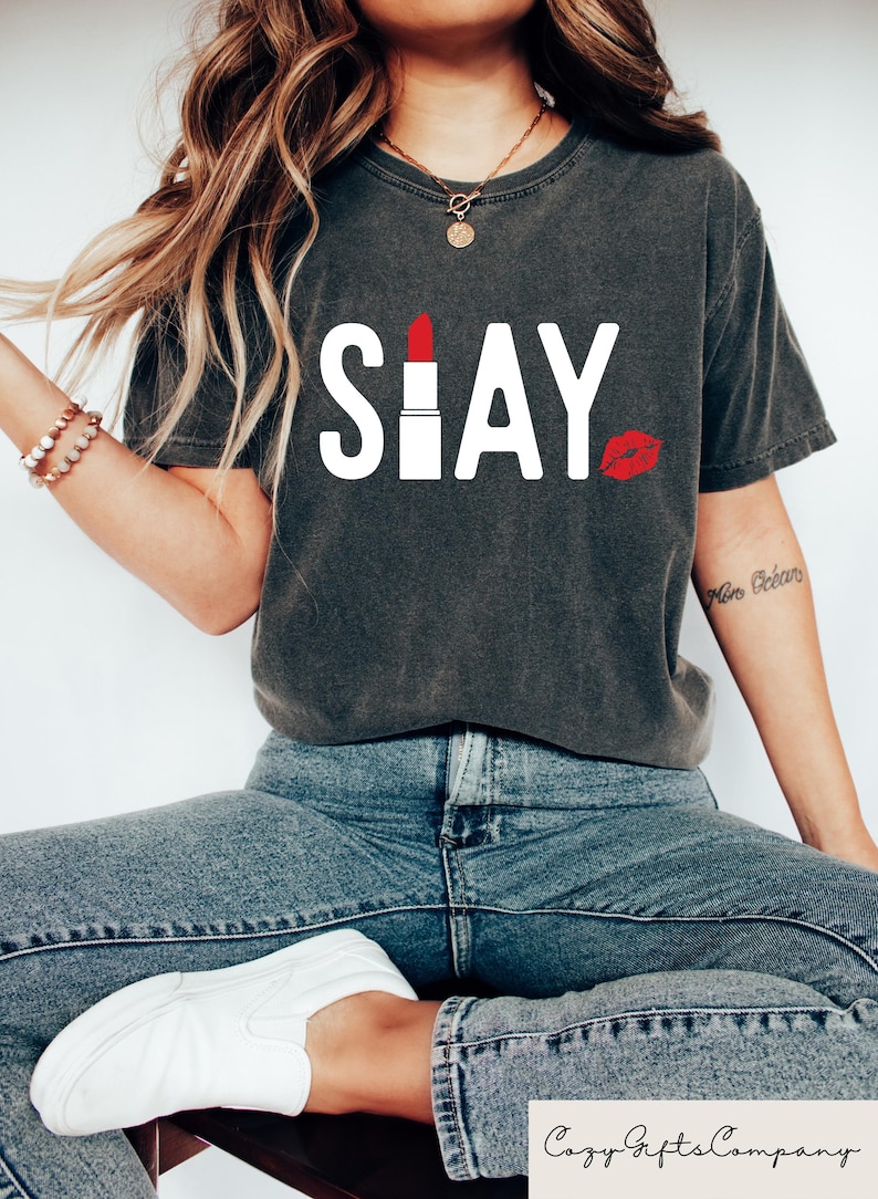 Puede incluir: Camiseta gris oscuro con la palabra "SLAY" en letras blancas. La "A" se reemplaza con un l&aacute;piz labial rojo, y una huella de labios roja est&aacute; a la derecha. La camiseta se combina con jeans azules y zapatillas blancas.
