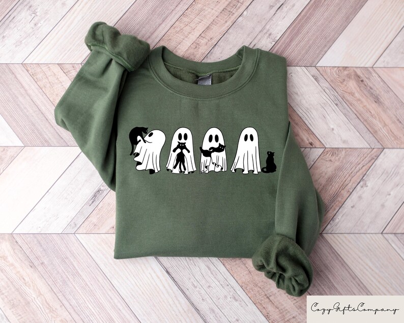 May include: A green sweatshirt with a graphic of five ghosts holding black cats. The ghosts are white and have black eyes and mouths. The cats are black and have white eyes.