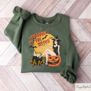 May include: Green sweatshirt with a graphic print of a witch sitting on a pumpkin. The text "Season of the Witch" is printed on the sweatshirt.