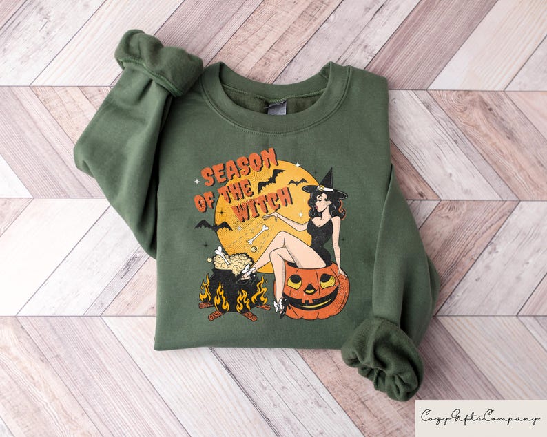 May include: Green sweatshirt with a graphic print of a witch sitting on a pumpkin. The text "Season of the Witch" is printed on the sweatshirt.