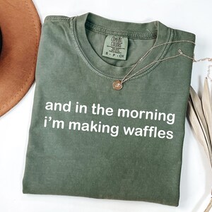 May include: Olive green t-shirt with the text "and in the morning i'm making waffles" in white. A gold necklace with a pendant is on the shirt. A brown hat and dried palm leaf are in the background.