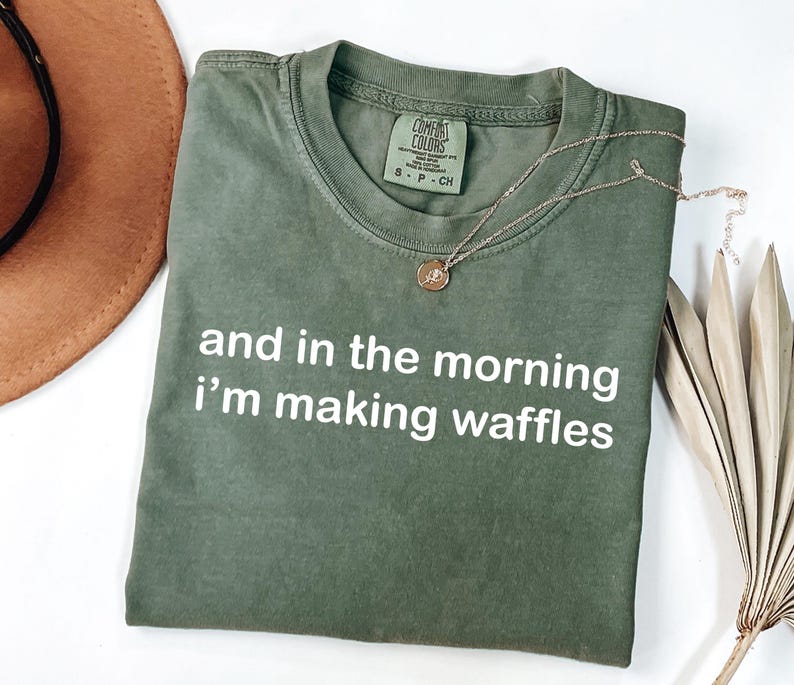 May include: Olive green t-shirt with the text "and in the morning i'm making waffles" in white. A gold necklace with a pendant is on the shirt. A brown hat and dried palm leaf are in the background.