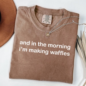 May include: A brown t-shirt with the text "and in the morning I'm making waffles" in white. A silver necklace with a pendant is draped across the shirt. The shirt is folded on a white surface.