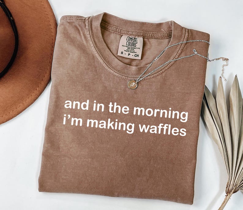 May include: A brown t-shirt with the text "and in the morning I'm making waffles" in white. A silver necklace with a pendant is draped across the shirt. The shirt is folded on a white surface.