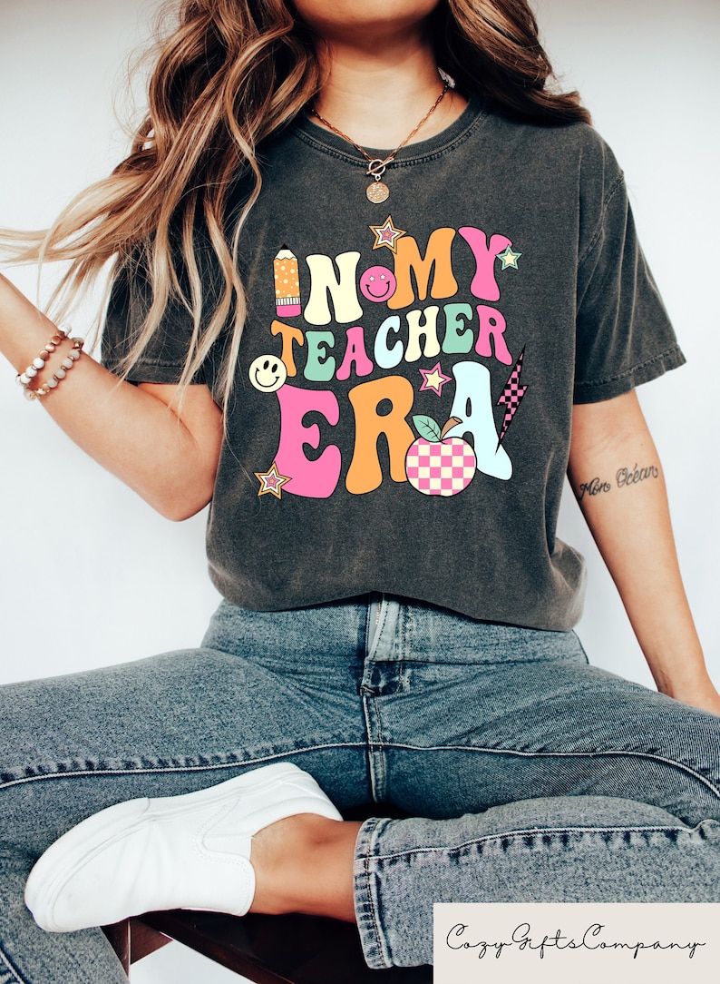 May include: Dark grey t-shirt featuring the text "IN MY TEACHER ERA" in a colourful, retro design. The design includes a pencil, smiley faces, stars, a lightning bolt, and a checkered apple. The shirt is worn with blue jeans and white trainers.